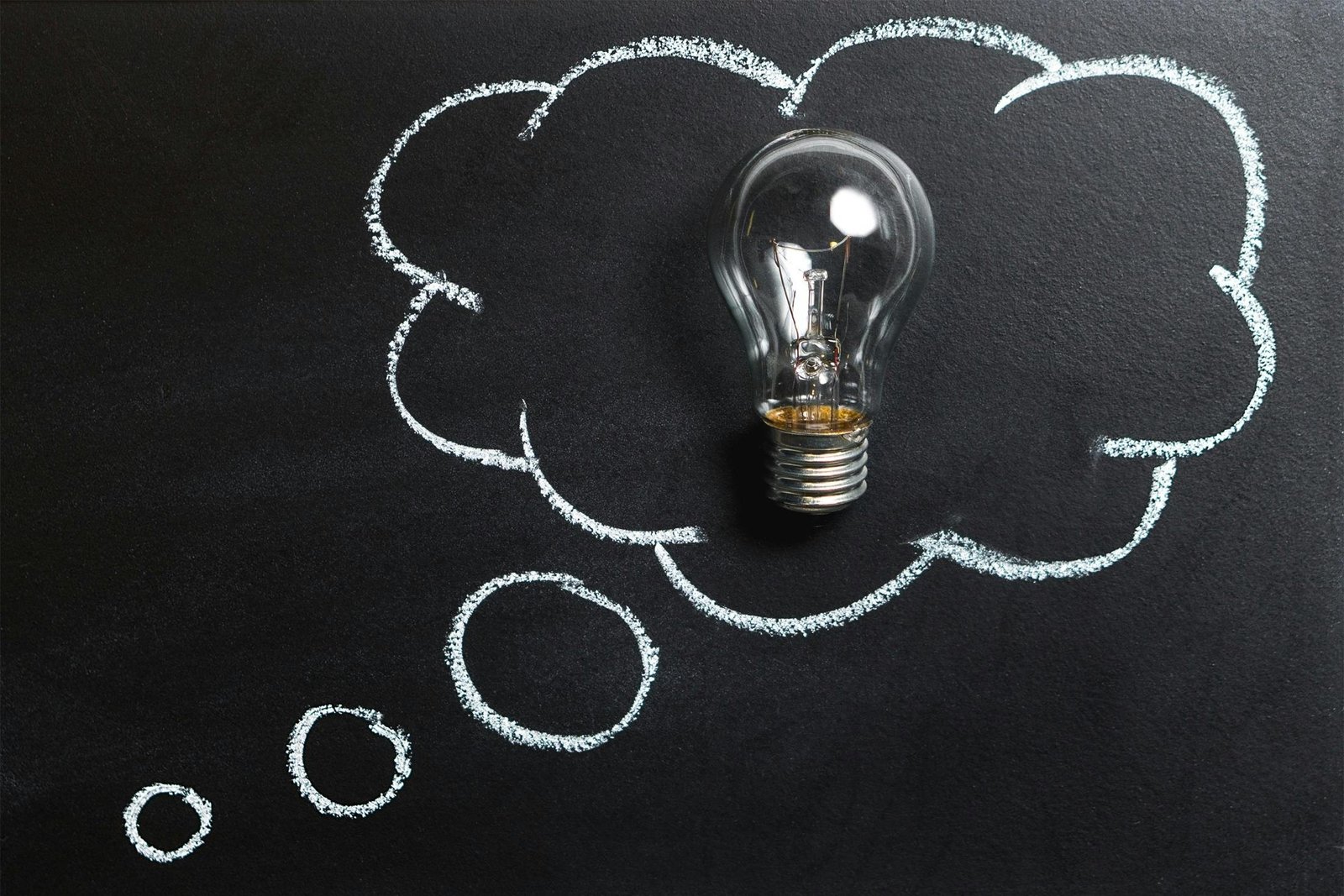 An incandescent lightbulb against a black background, with a cloud drawn around it in chalk