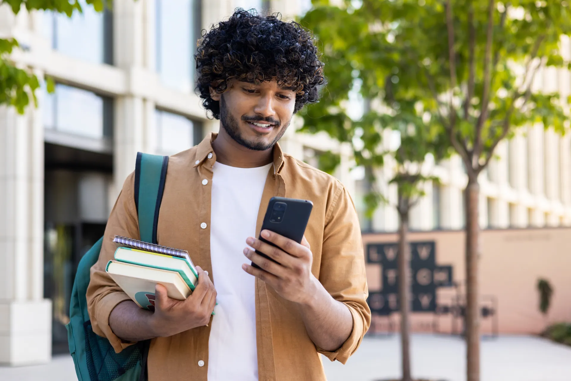 A confident-looking student leaving their language school. They are carrying textbooks and looking down at their smartphone.