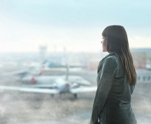 A woman standing inside an airport, looking out through a window to planes on the tarmac.